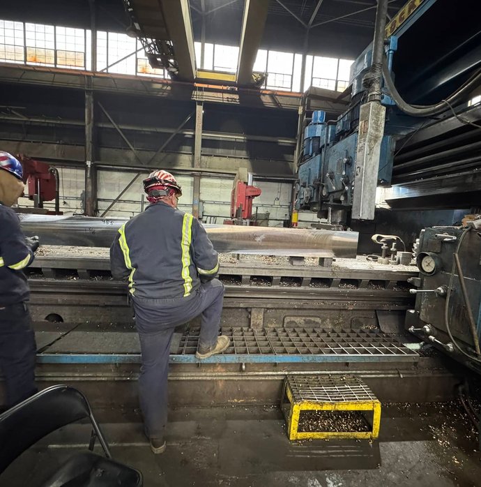 Two workers in safety gear inspecting a freshly machined steel shaft